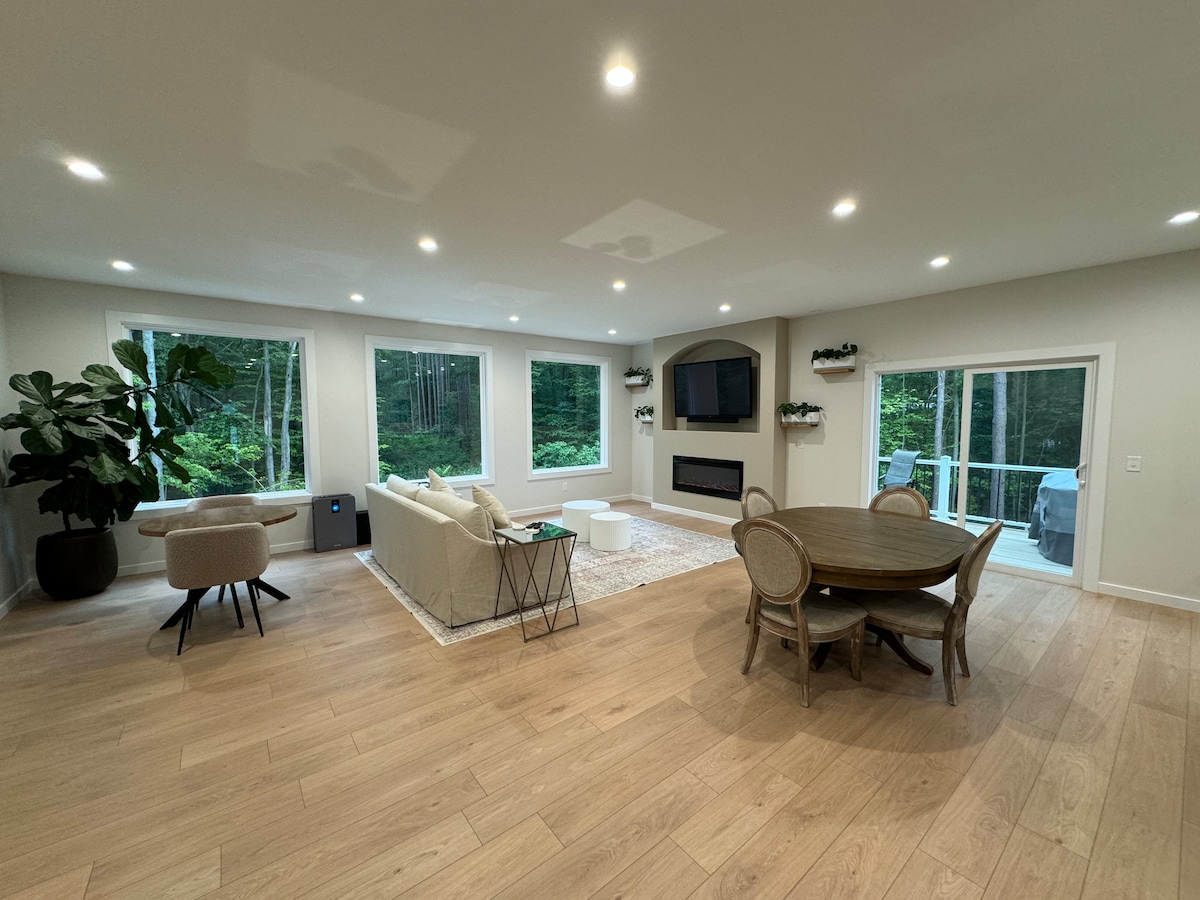The spacious living area features large windows that frame a view of the surrounding greenery. A comfortable sofa is positioned near a sleek fireplace, while a round dining table with wicker chairs is situated nearby. Warm wood flooring adds to the serene atmosphere.