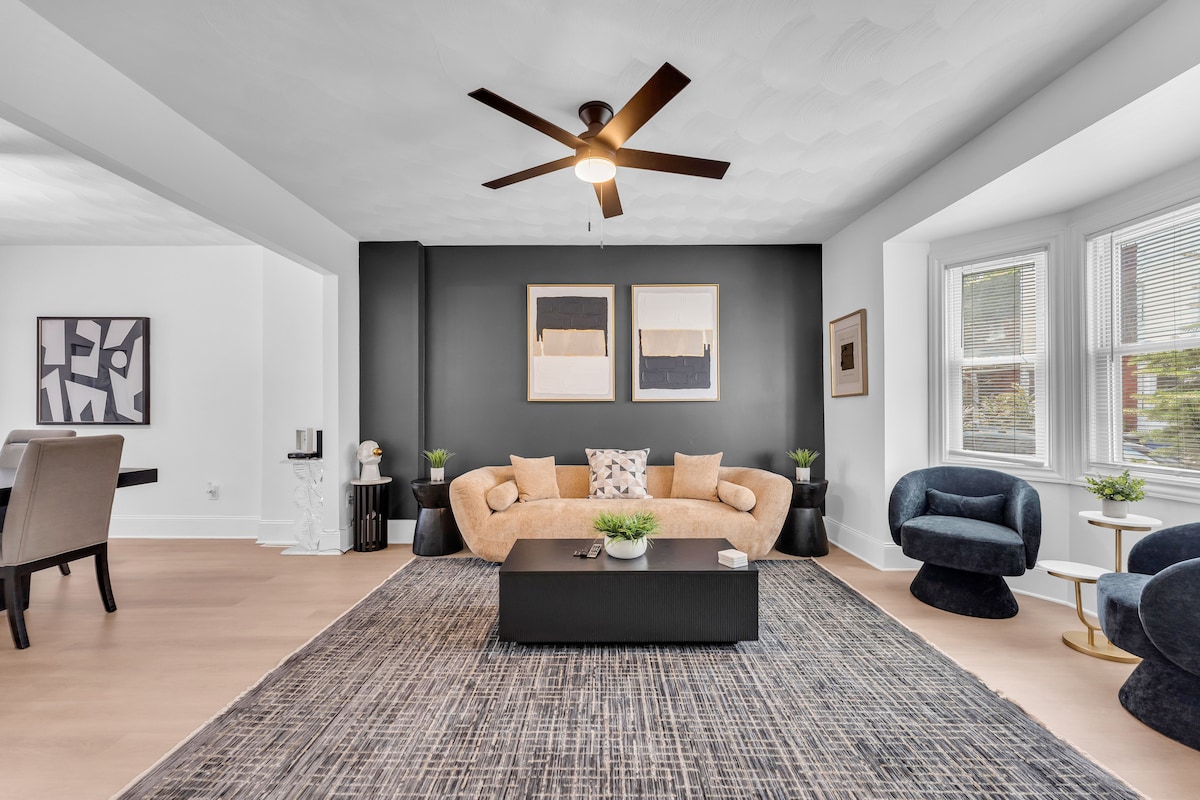 An open living area features a large cream-colored sofa arranged around a dark coffee table, complemented by two dark plush chairs. A fan hangs above, and natural light enters through multiple windows. Simple artwork is displayed on the black accent wall, enhancing the modern aesthetic.