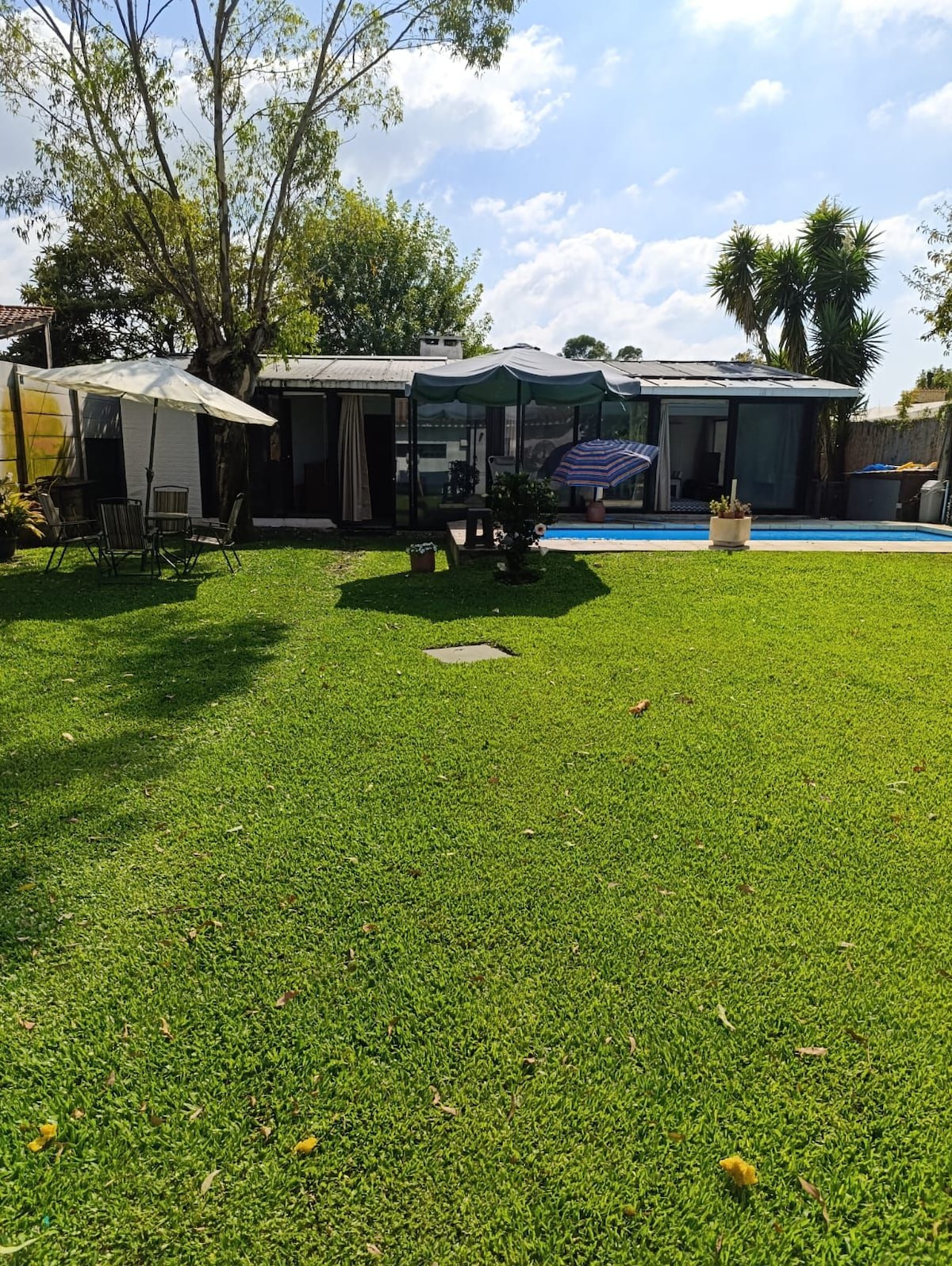 A spacious garden area features lush green grass, with a swimming pool reflecting sunlight. Comfortable seating is arranged beneath a shaded gazebo. The rear of the house, characterized by large glass doors, opens up to the garden, providing a seamless transition between indoor and outdoor spaces.