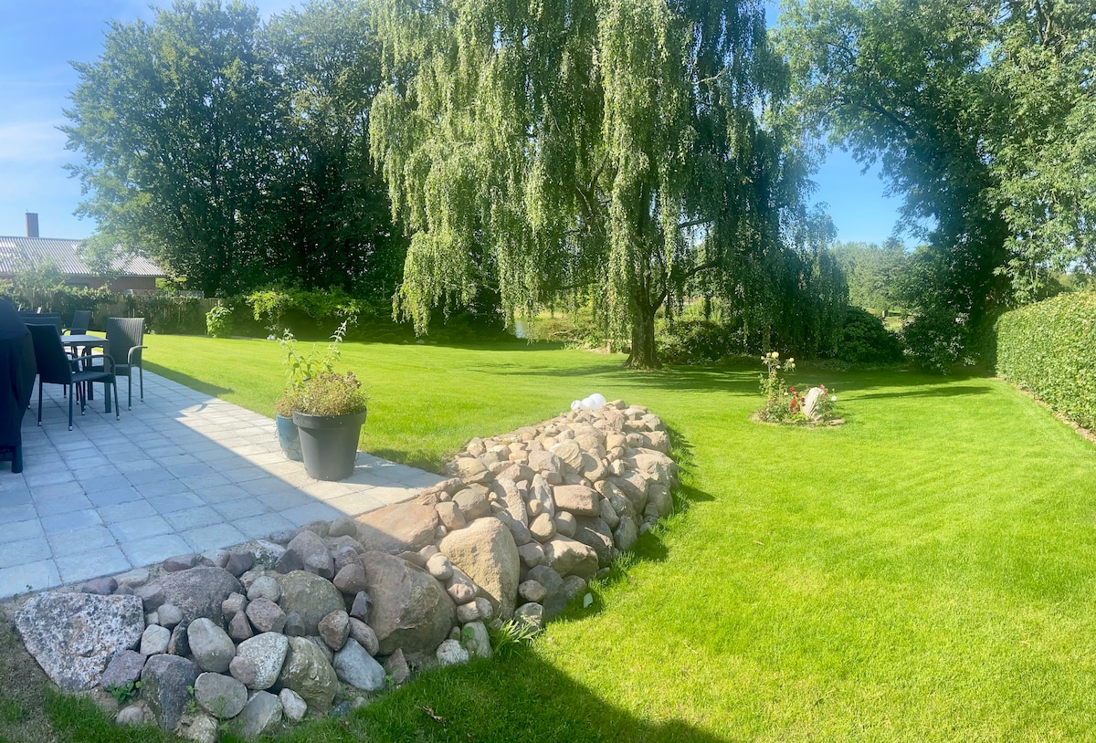 A lush garden area features well-maintained green grass, bordered by a pebble rock formation. A patio with black outdoor furniture is visible, providing a space for relaxation. Mature trees provide shade, and flowering plants enhance the natural setting, contributing to a serene outdoor environment.