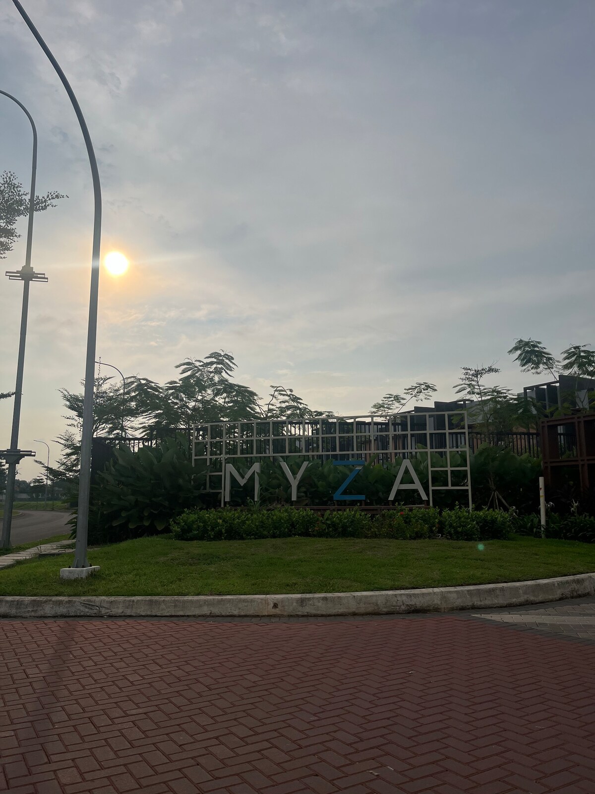 The entrance sign of Myza BSD is prominently displayed amidst lush greenery. The sign features large, modern letters with 'MYZA' highlighted in blue. A soft sunlight casts a gentle glow over the surroundings, contributing to a welcoming atmosphere.