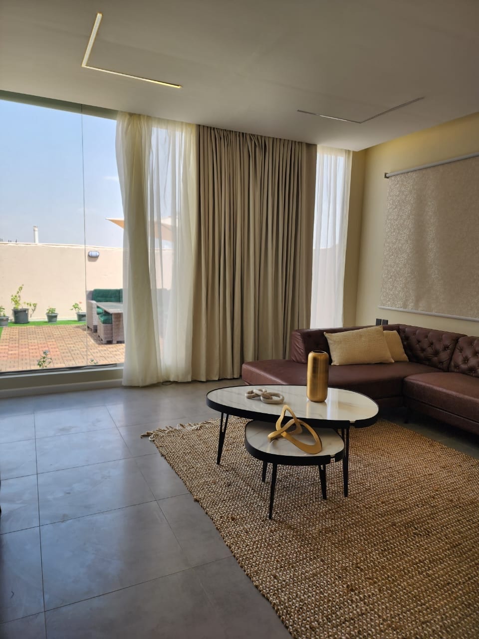 A spacious living area features a large window offering natural light and views of a private terrace. A comfortable brown couch is positioned opposite a round coffee table, while a textured beige rug lays underfoot, enhancing the room's modern aesthetic.