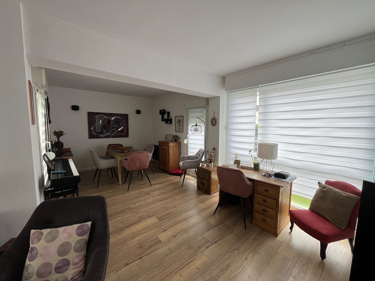 A cozy common area features wooden flooring with various seating options, including a desk and armchairs. Natural light filters in through large windows dressed in light blinds. A piano and decorative elements create a warm and functional space for relaxation and socializing.