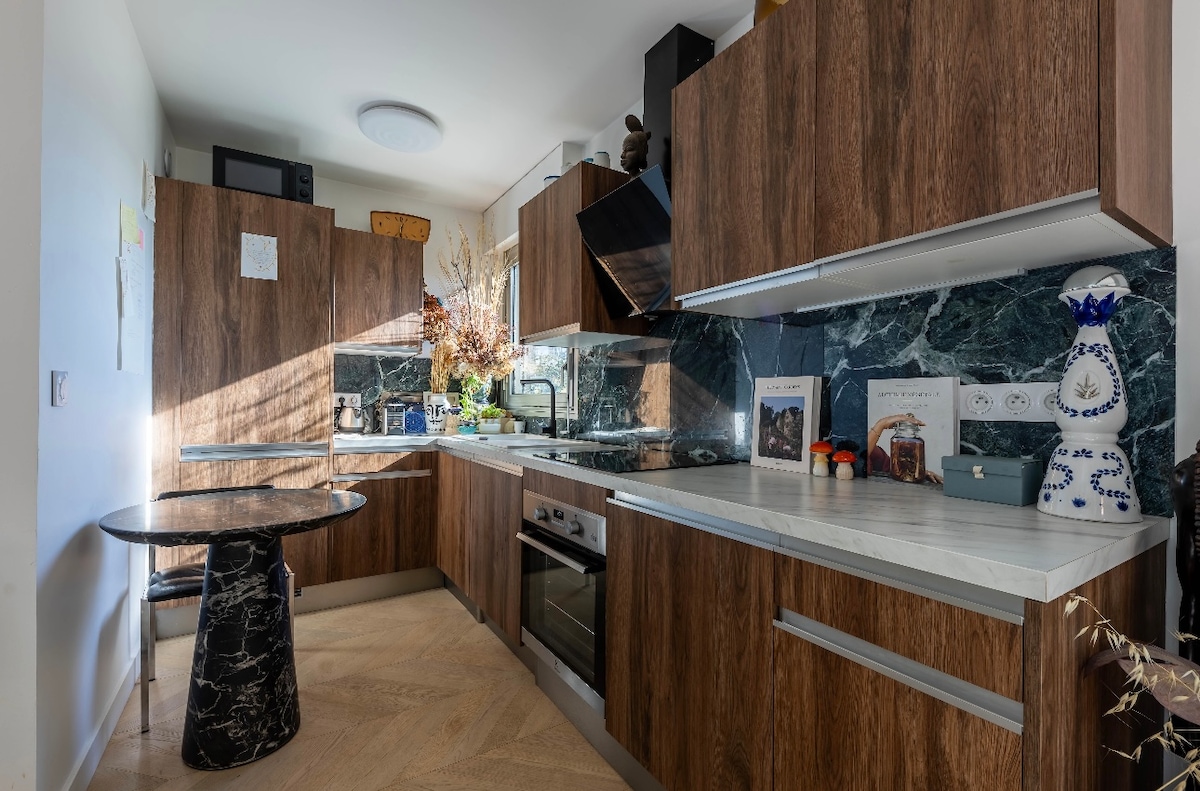 The kitchen features wooden cabinetry with a modern design, complemented by a marble countertop. A round table with a dark stone base is positioned in the corner. Natural light streams in, illuminating the sleek appliances and decorative items arranged on the shelves.