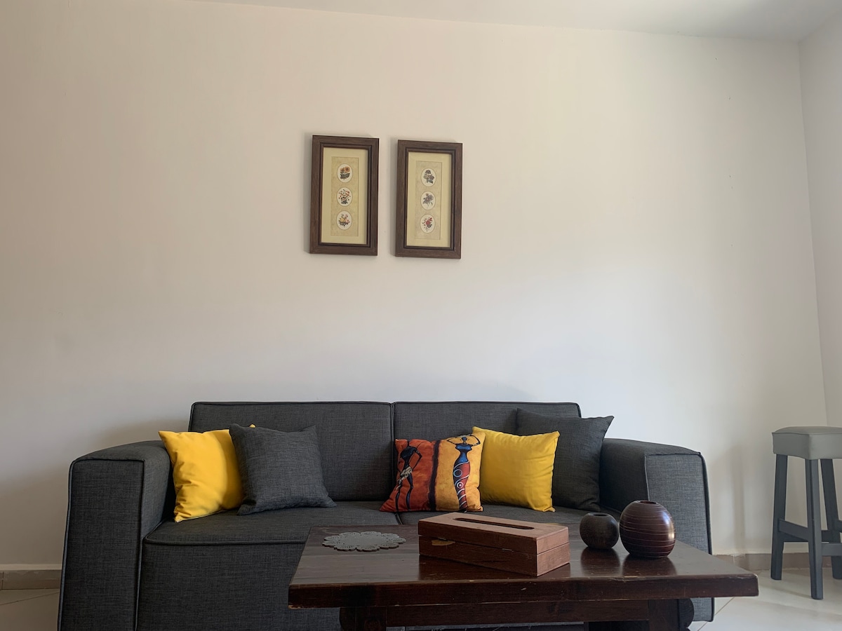A modern living area features a gray sofa adorned with colorful cushions in yellow and gray. A wooden coffee table with decorative items sits in front of the sofa. Two framed artworks are displayed on the white wall above the sofa.