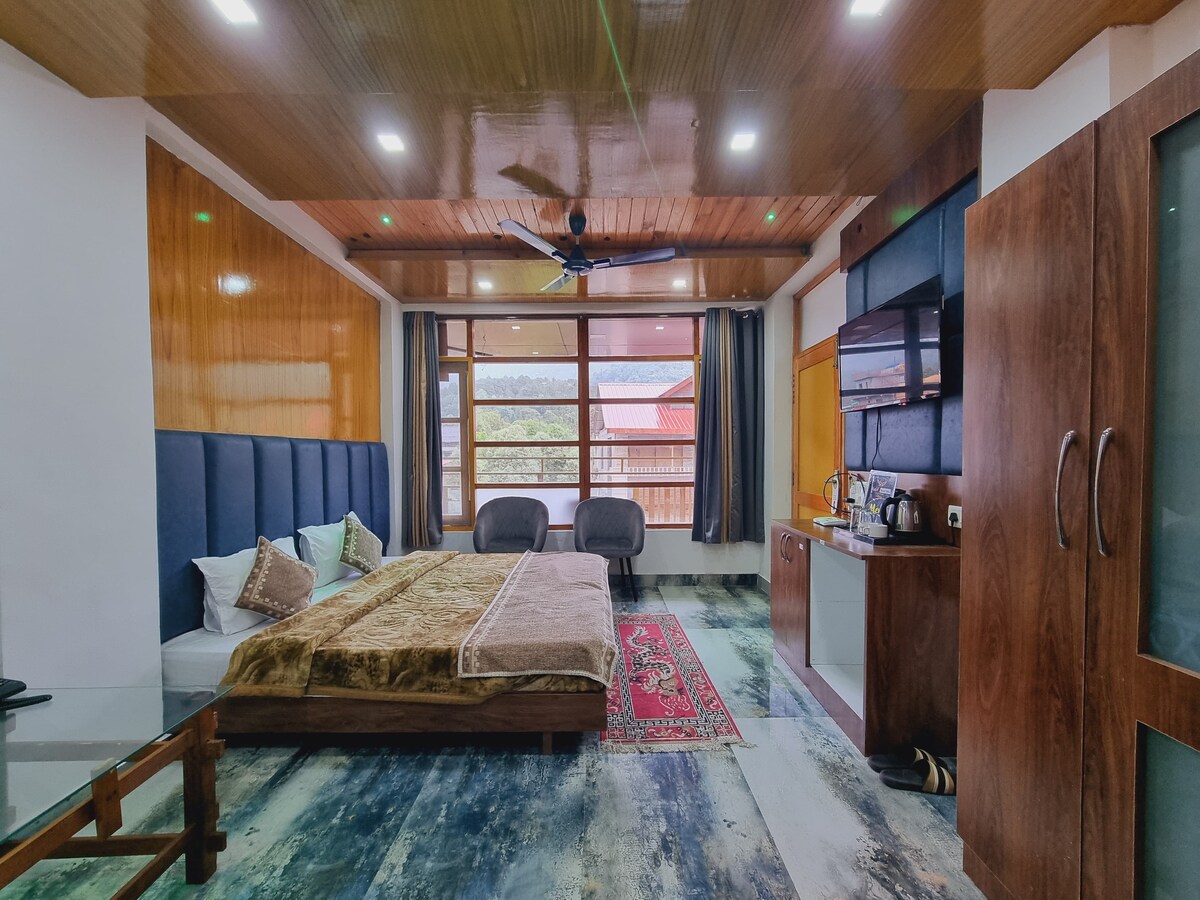 A spacious room features a large bed with a decorative blanket, accompanied by two armchairs. Natural light is allowed through large windows with scenic views. The room contains a kitchenette, and wooden elements add warmth to the atmosphere.