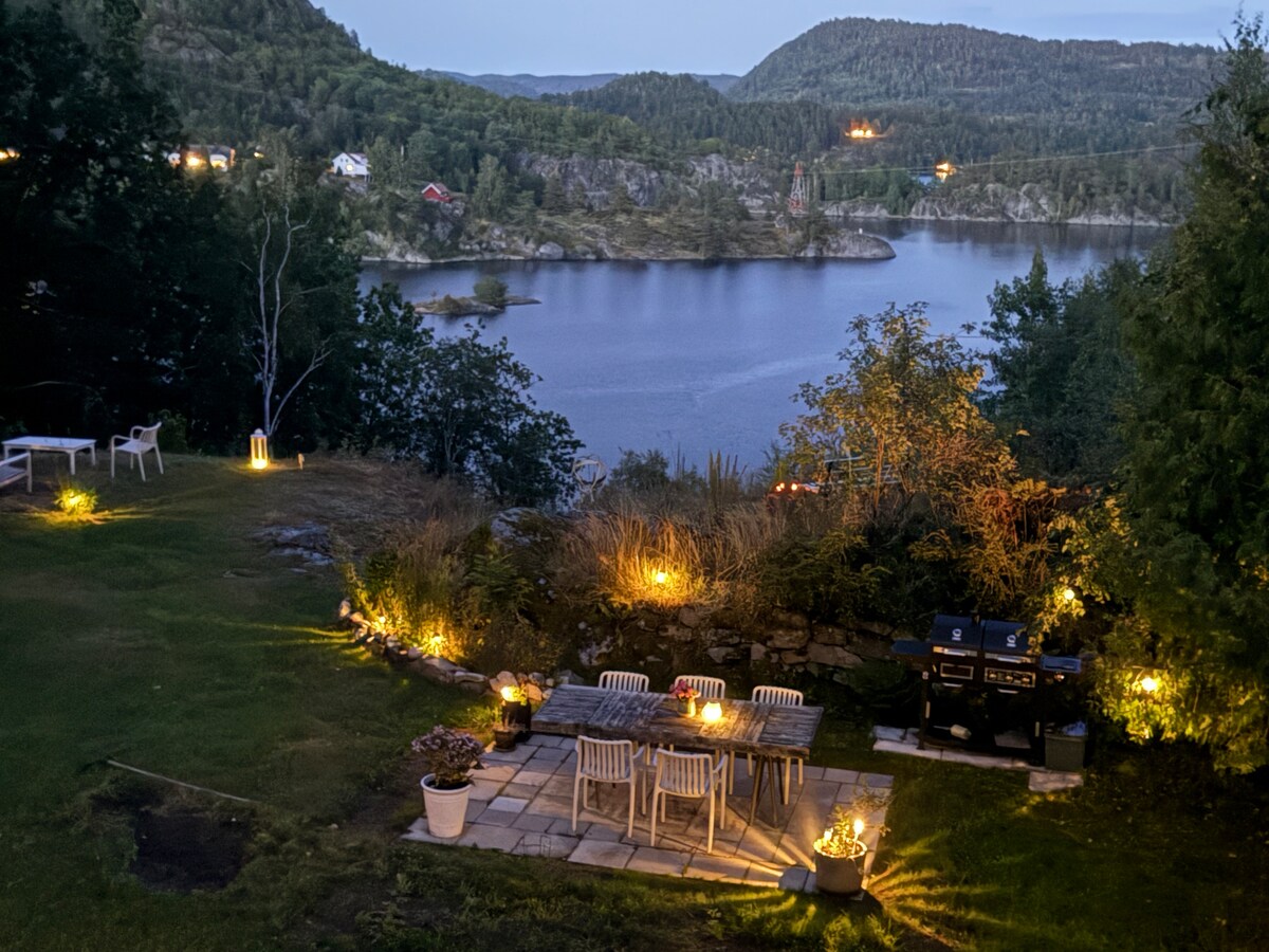 An outdoor patio area is illuminated with soft lighting, featuring a table surrounded by chairs. A barbecue grill is present nearby. Lush greenery contrasts with the calm water of the fjord in the background, creating a serene atmosphere at dusk.