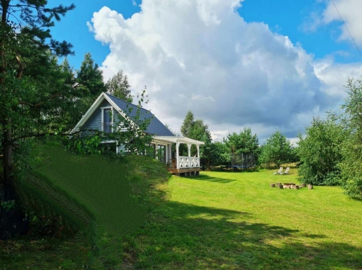 A house with a covered porch is set amidst expansive green lawns and trees. The sky features clouds casting dynamic shadows across the property, and a small seating area made of logs is visible in the background, inviting outdoor relaxation.