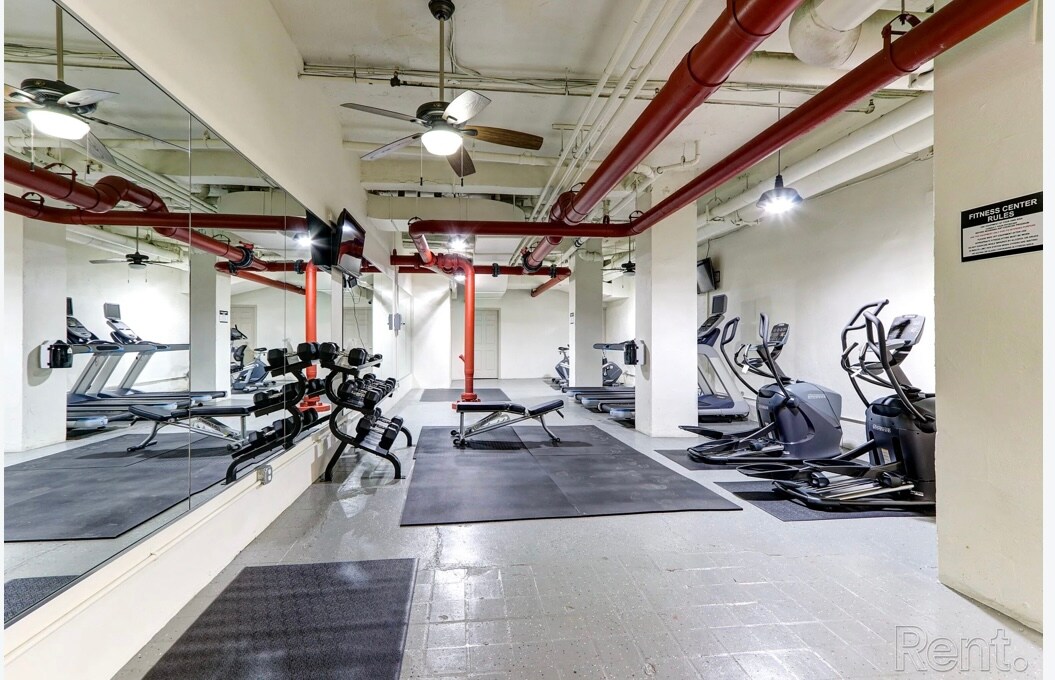 A fitness room is equipped with modern workout machines including treadmills, elliptical trainers, and free weights. The space features large mirrors on one wall and is complemented by overhead lighting and ventilation fans, creating a spacious and functional exercise environment.