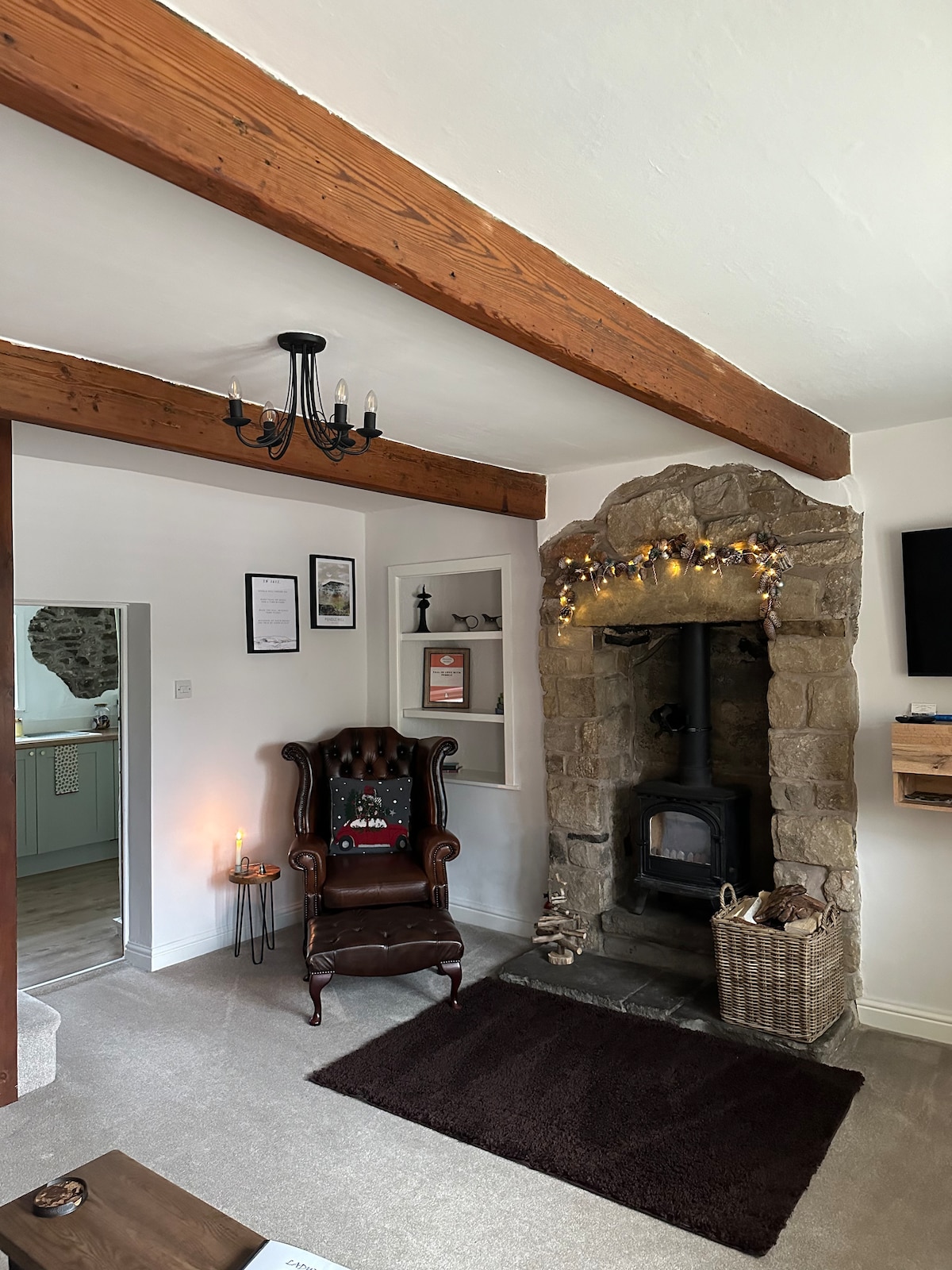 A cozy living area features exposed wooden beams and a stone fireplace with a log burner. A vintage-style armchair is positioned beside a soft rug. Shelves display decorative items, while gentle lighting enhances the warm ambiance, creating an inviting retreat.