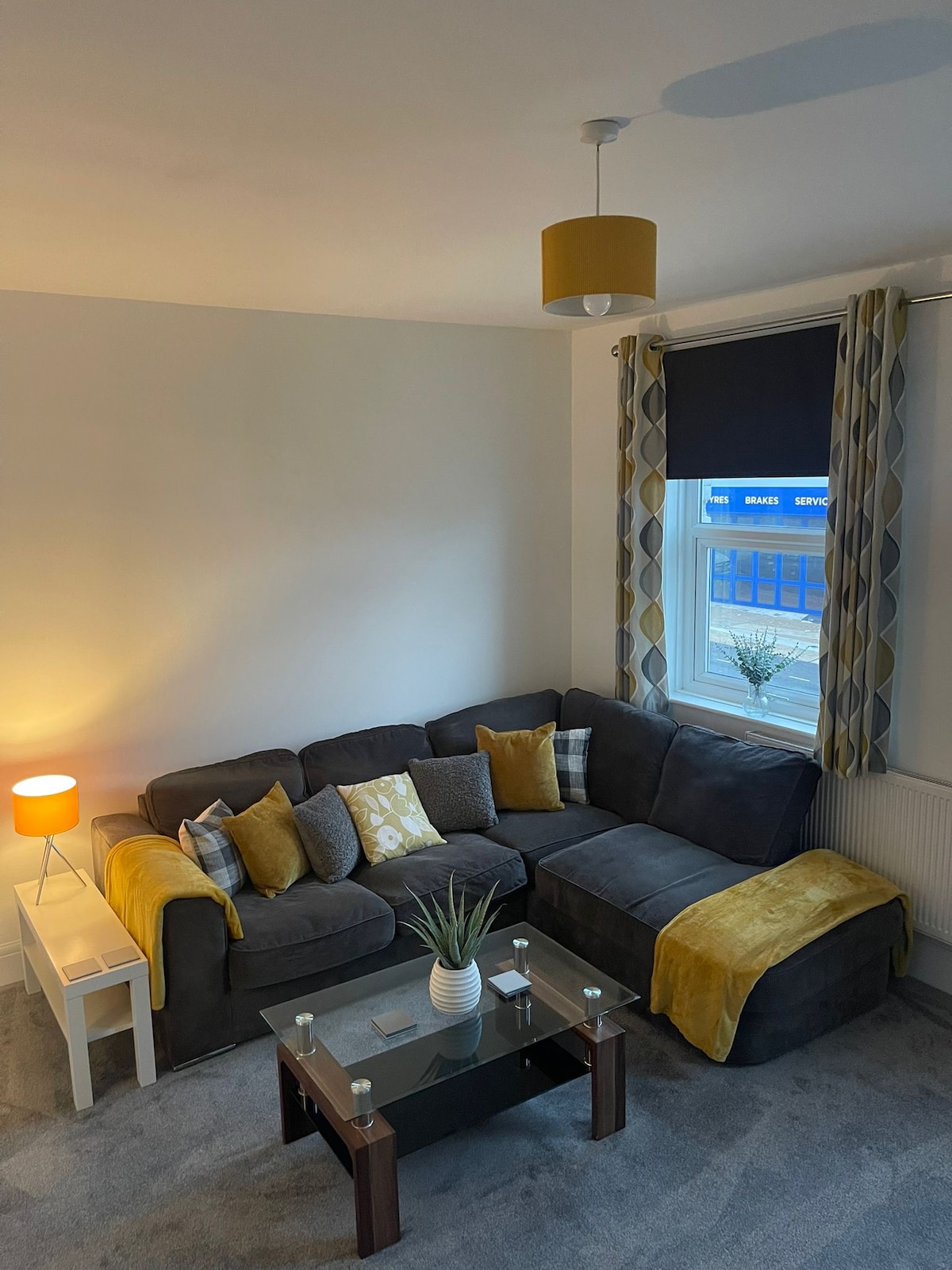 A modern living area is presented with a dark sectional sofa adorned with a variety of cushions in soft hues. A sleek glass coffee table sits in front, complemented by a decorative plant. Light is provided by a floor lamp and window treatments filter external light.