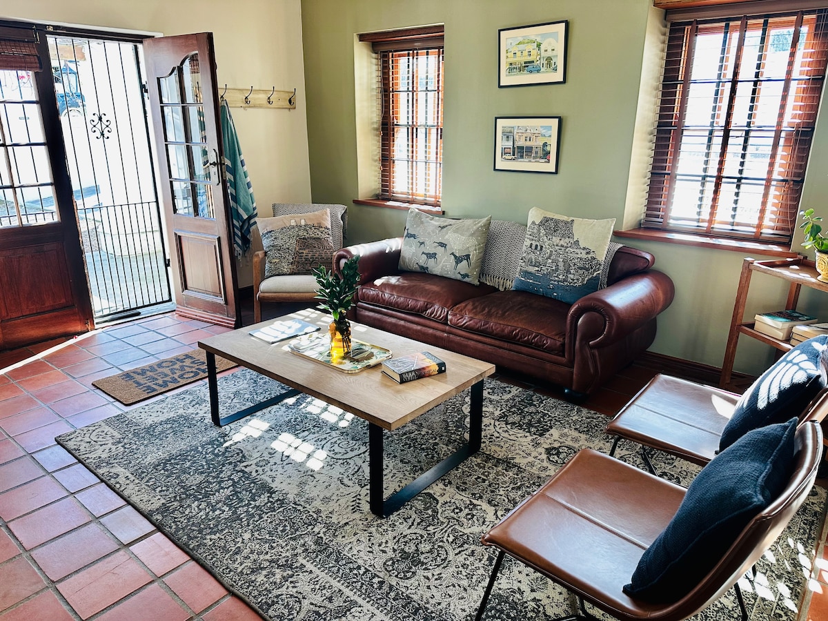 A comfortable living area is presented with a brown leather sofa adorned with decorative pillows. Two stylish chairs are arranged nearby, accompanied by a coffee table displaying books. Light filters in through wooden window blinds, creating a warm ambiance, while a welcoming entrance is visible.