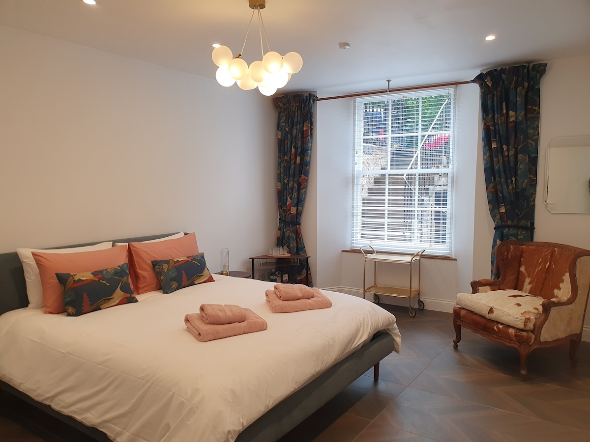 A spacious bedroom features a superking bed with decorative pillows. A stylish sitting chair complements the decor, and a drinks station is available for convenience. Natural light filters through a large window adorned with patterned curtains, illuminating the warm tones of the furnishings.