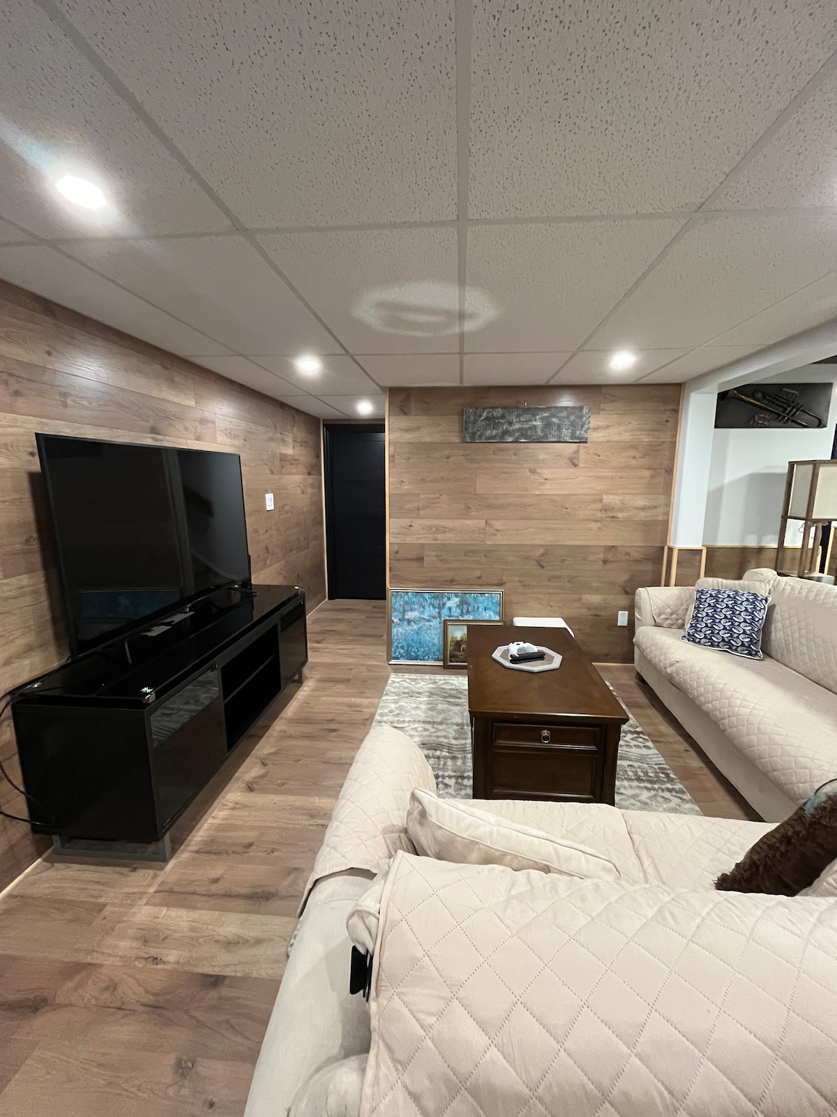 A modern living area features a large flat-screen TV mounted on a wood-paneled wall, complemented by a stylish coffee table. Light-colored couches are arranged to create a cozy seating arrangement, with decorative pillows adding subtle textures. A plush area rug lies beneath, enhancing the inviting atmosphere.