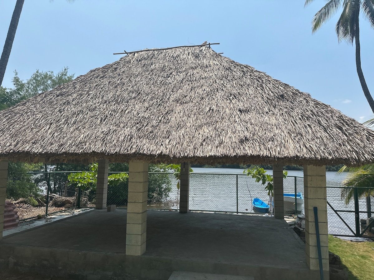 A thatched-roof gazebo supported by concrete pillars is viewed from the front. The structure overlooks a serene waterway, with green foliage and a fence surrounding the area. A small glimpse of blue boats is visible in the calm water beyond the gazebo.