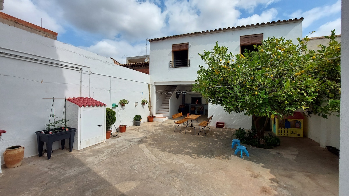 The spacious outdoor patio is surrounded by white walls, featuring a large tree, tables, and chairs. A small playhouse and a storage shed with a red roof are visible, adding functionality to the area. The sky is partly cloudy, creating a serene atmosphere.