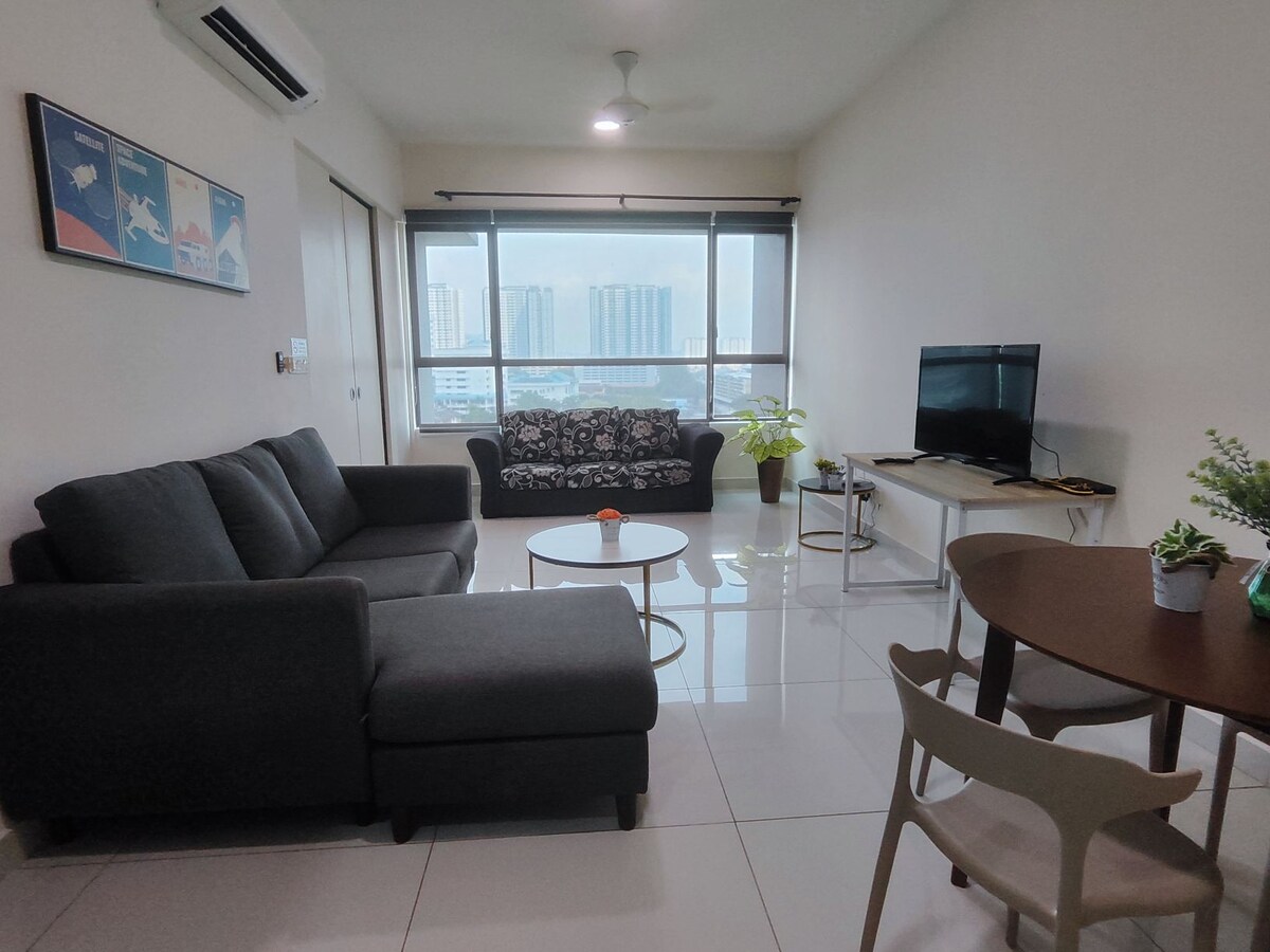 A modern living area features a comfortable couch and armchair, with a round coffee table positioned at the center. Large windows provide ample natural light, revealing a view of the surrounding buildings. A television is placed on a stand, complemented by a decorative plant.