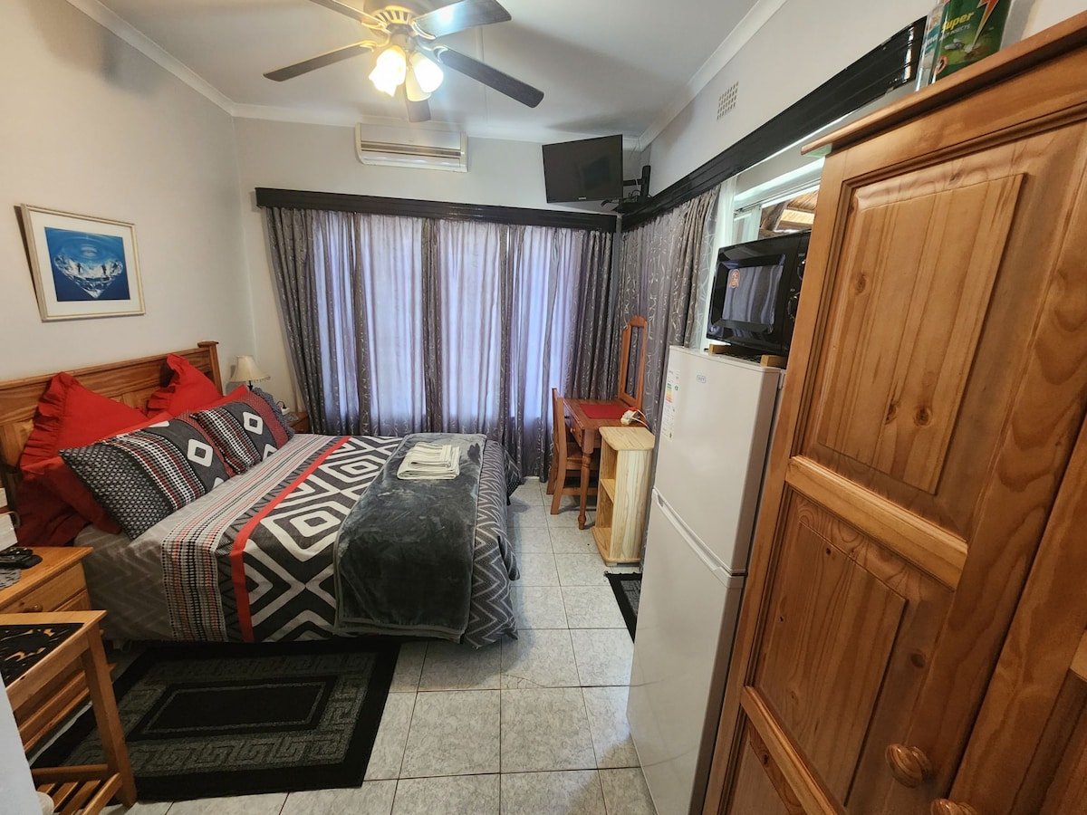 A compact room features a spacious bed adorned with red and gray bedding. Wooden furniture, including a storage cabinet and a small desk, creates a functional space. Natural light filters through the partially closed curtains, and a ceiling fan provides additional comfort.