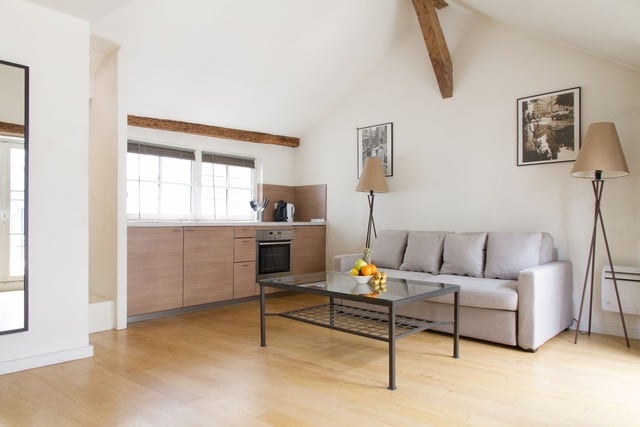 A bright and airy living space features a modern kitchenette with wooden cabinetry and sleek appliances. A comfortable sofa sits across from a glass coffee table, complemented by two elegant floor lamps. Natural light floods the room through large windows, enhancing the clean lines of the decor.
