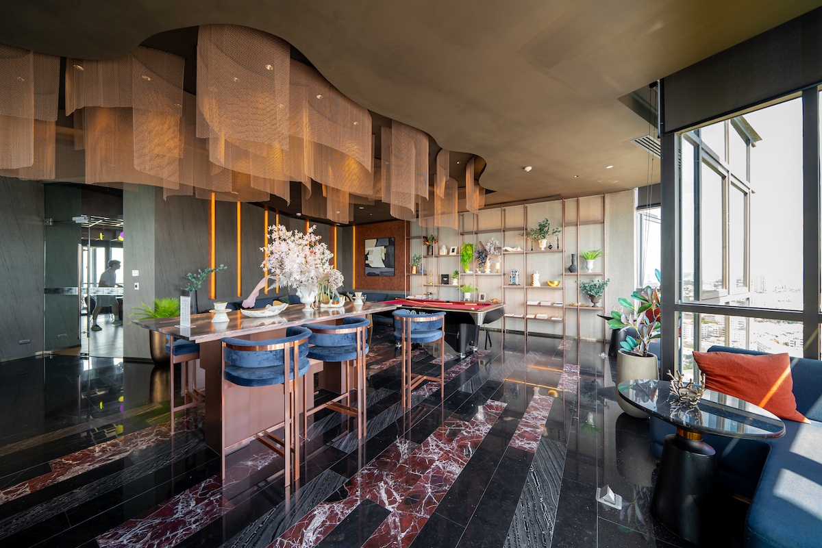 An open-plan dining area features a large table surrounded by comfortable chairs. The space is complemented by elegant light fixtures and sleek black marble flooring, while various plants and decorative elements add a touch of greenery to the room.