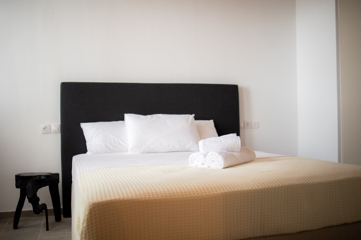 A minimalist sleeping area is featured, with a spacious bed dressed in neutral tones and topped with white linens. Soft towels are neatly arranged at the foot of the bed. A small, dark accent table enhances the simple design.