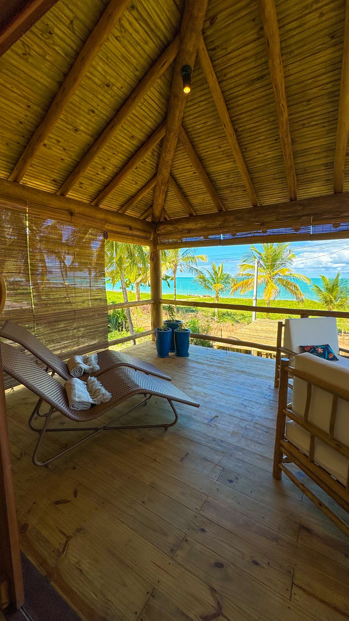 A sunlit balcony area features a large lounging chair and a small wooden table. The natural bamboo ceiling offers a rustic touch, while expansive views of the ocean and palm trees can be seen through the open space, creating a serene environment.