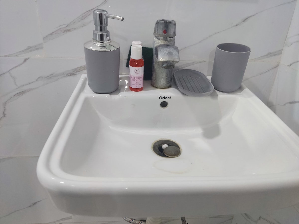 A clean, modern sink area is displayed with a sleek soap dispenser and a small bottle on the countertop. A matching cup complements the aesthetic. The sink features a polished faucet, surrounded by a light-colored tiled wall, providing a fresh and functional space.