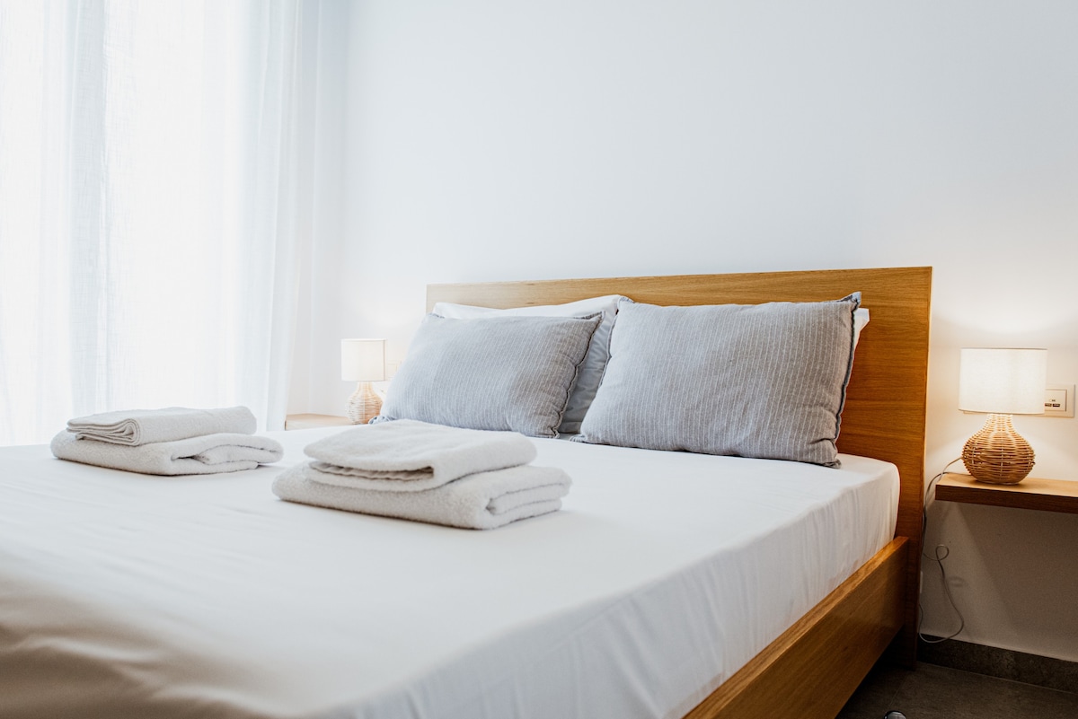 A spacious bed is centered in the room, featuring soft grey pillows and neatly folded white towels placed at the foot. Warm bedside lamps are positioned on minimalist nightstands, offering gentle illumination. Natural light filters through sheer curtains, enhancing the serene atmosphere.