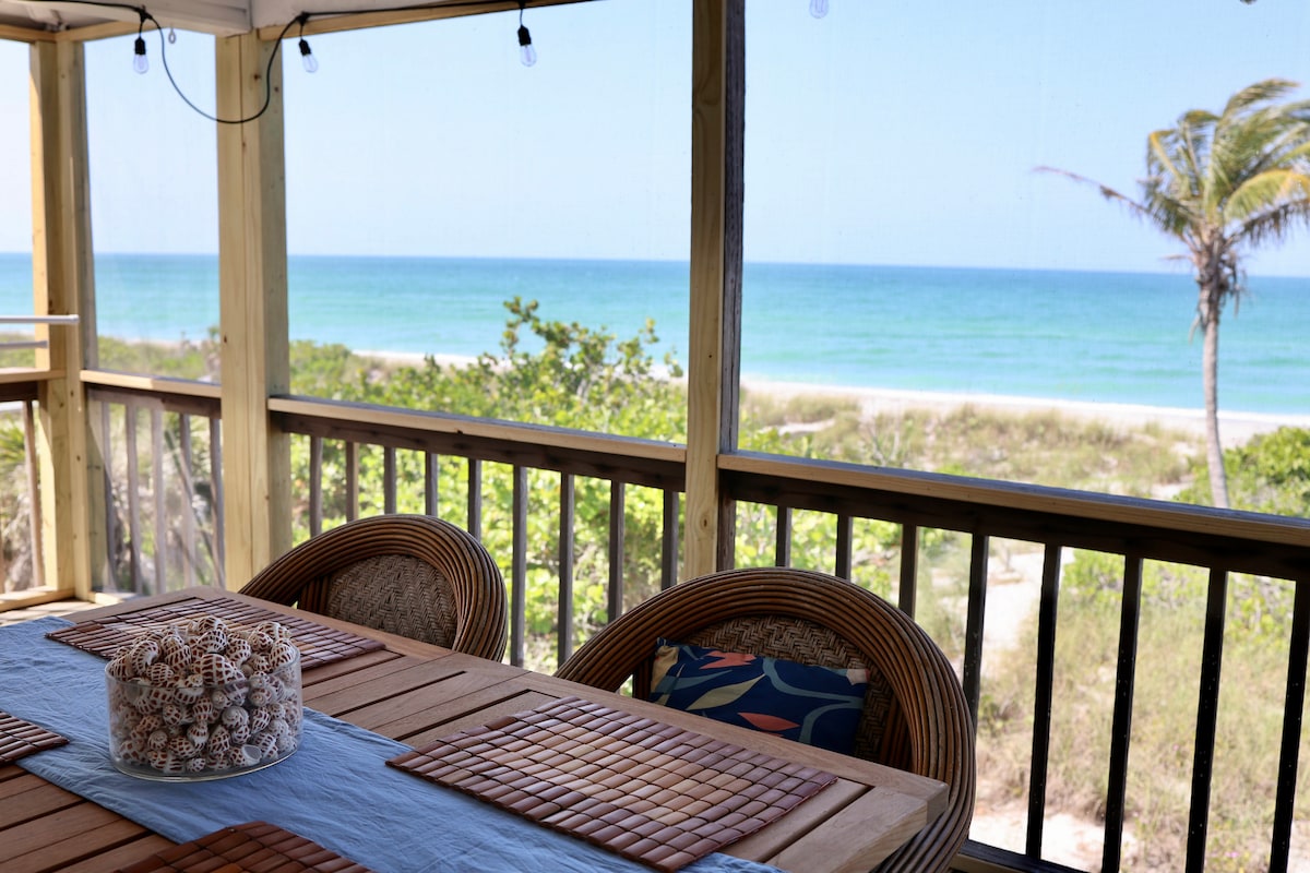 A dining area is positioned on a screened porch, featuring a wooden table surrounded by four chairs. A decorative bowl filled with shells rests on the table. Beyond the railing, scenic beach views and calm turquoise waters are visible, enhancing the space's relaxed atmosphere.