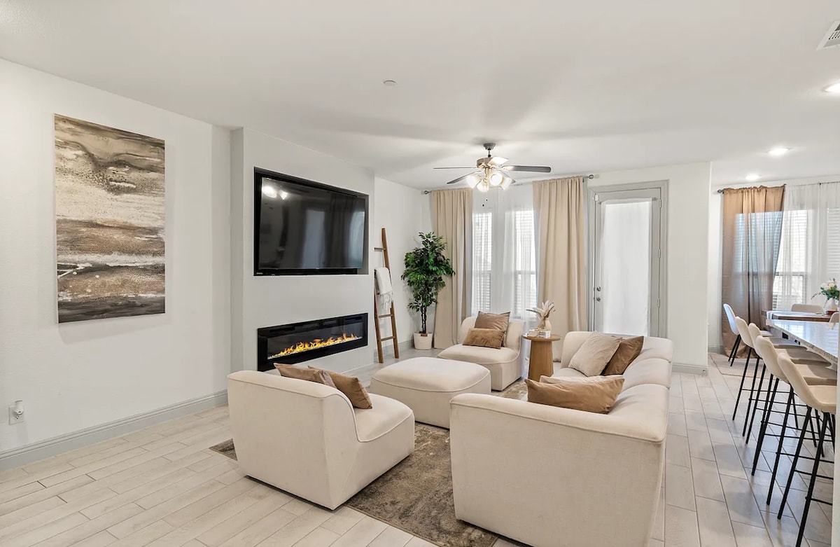 A comfortable living room features a sectional sofa and two armchairs, accented with neutral pillows. A wall-mounted smart TV and an electric fireplace are prominent. Windows adorned with light curtains allow natural light to fill the space, complemented by an area rug underfoot.