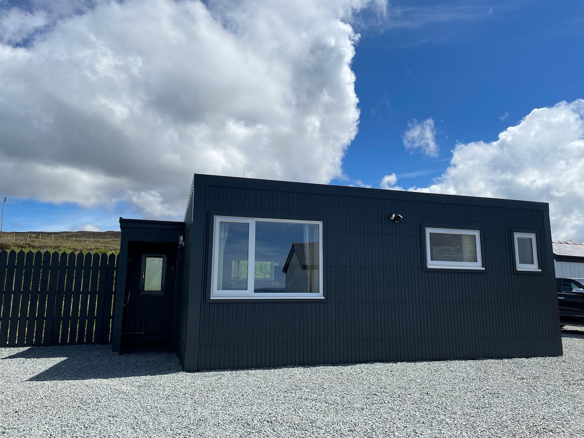 The exterior of a dark grey cabin is presented, featuring large windows that offer a glimpse of the surrounding landscape. A small porch leads to the entrance, with a fenced area enhancing the privacy. The sky is partly cloudy, creating a bright atmosphere.