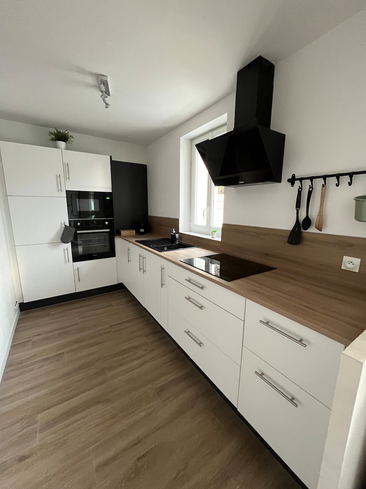 A modern kitchen is presented, featuring crisp white cabinets with sleek handles. A dark hood hangs above a polished countertop, complemented by a built-in oven and microwave. Natural light streams through a window, illuminating the well-organized space equipped for cooking.