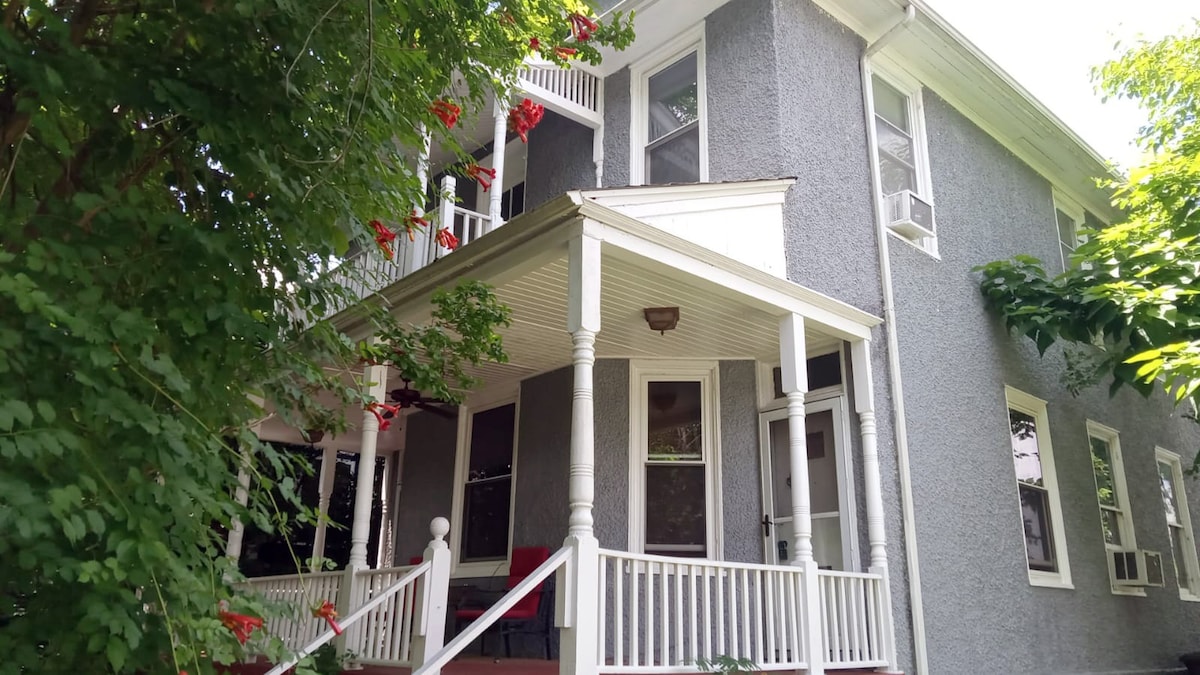A charming exterior of a two-story gray house is framed by lush greenery. A welcoming porch with white railings invites guests, featuring potted red flowers that add a pop of color. Windows allow ample natural light, enhancing the inviting facade.
