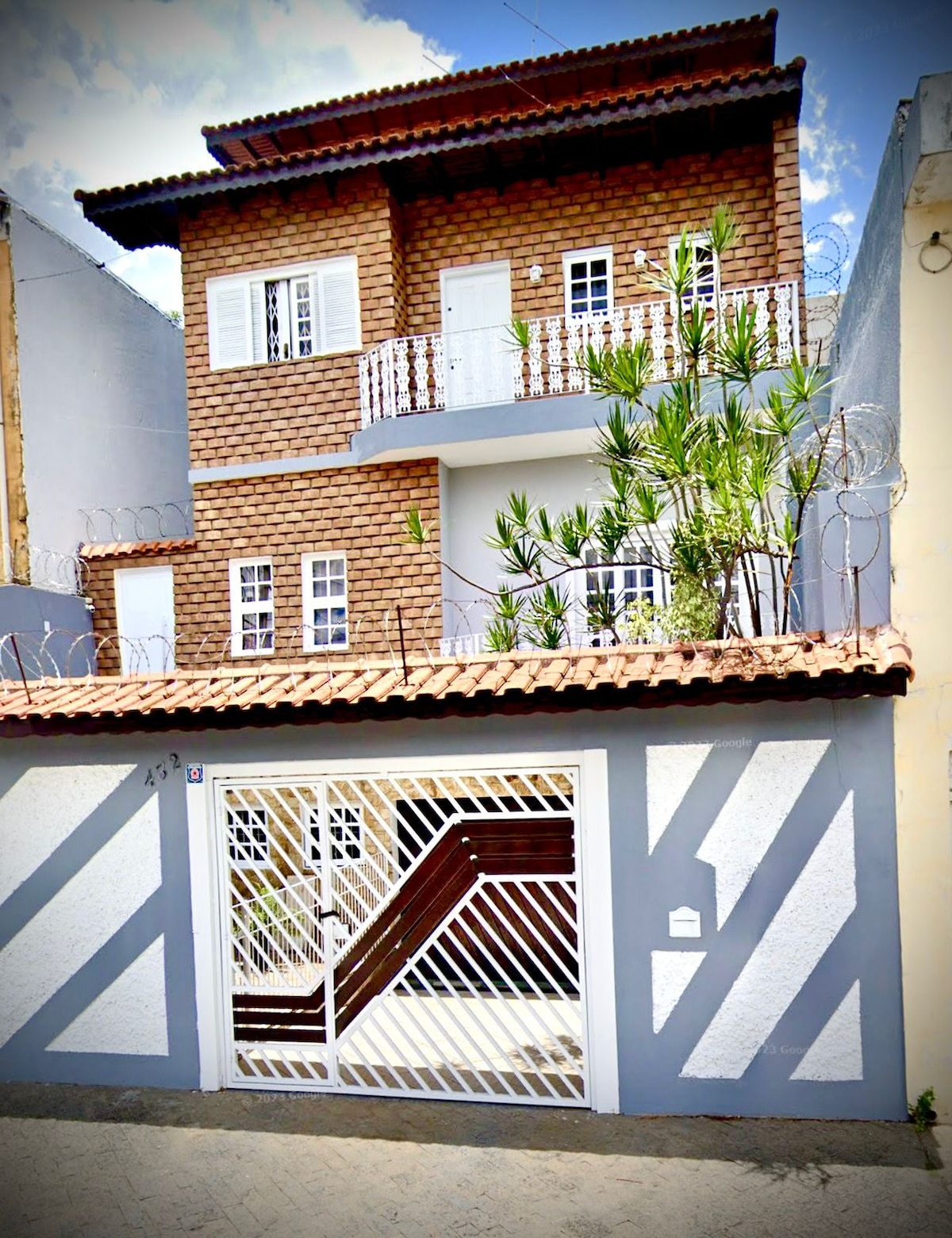 A three-story building with a textured brick facade is visible, featuring multiple windows framed in white. A tiled roof with overhang shades the entrance, which is adorned with plants. A gated entry allows access, complemented by a staircase leading to the main door.