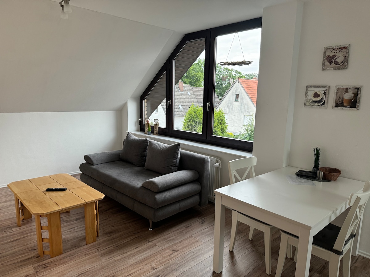 A bright living area features a dark gray sofa facing large windows that provide ample natural light. A wooden coffee table sits in front, while a white dining table and chairs are positioned nearby. Decorative wall art and a plant enhance the space.