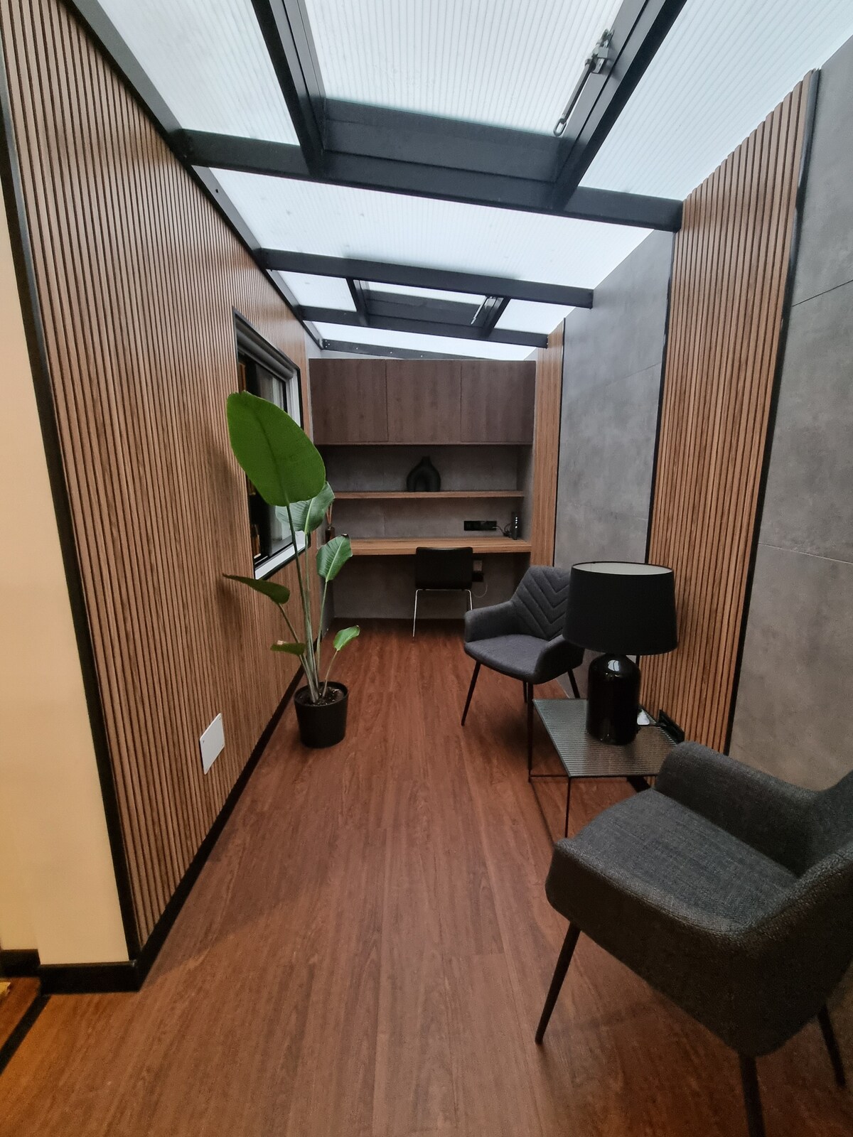 A modern hallway features vertical wooden paneling and gray concrete walls. A small desk is positioned against the back wall, accompanied by a potted plant and a lamp on a side table. Two chairs are placed along the path, creating a functional and aesthetically pleasing workspace.