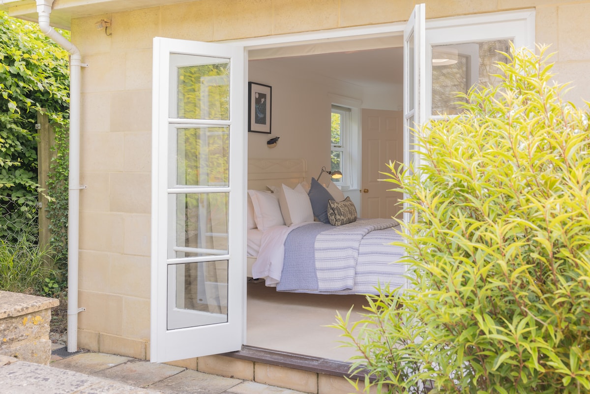 A light-filled bedroom is visible through open French doors, framed by lush greenery. The inviting space features a spacious bed adorned with soft bedding and decorative pillows, complemented by a bedside lamp and artwork on the wall. Soft carpeting extends throughout the room.