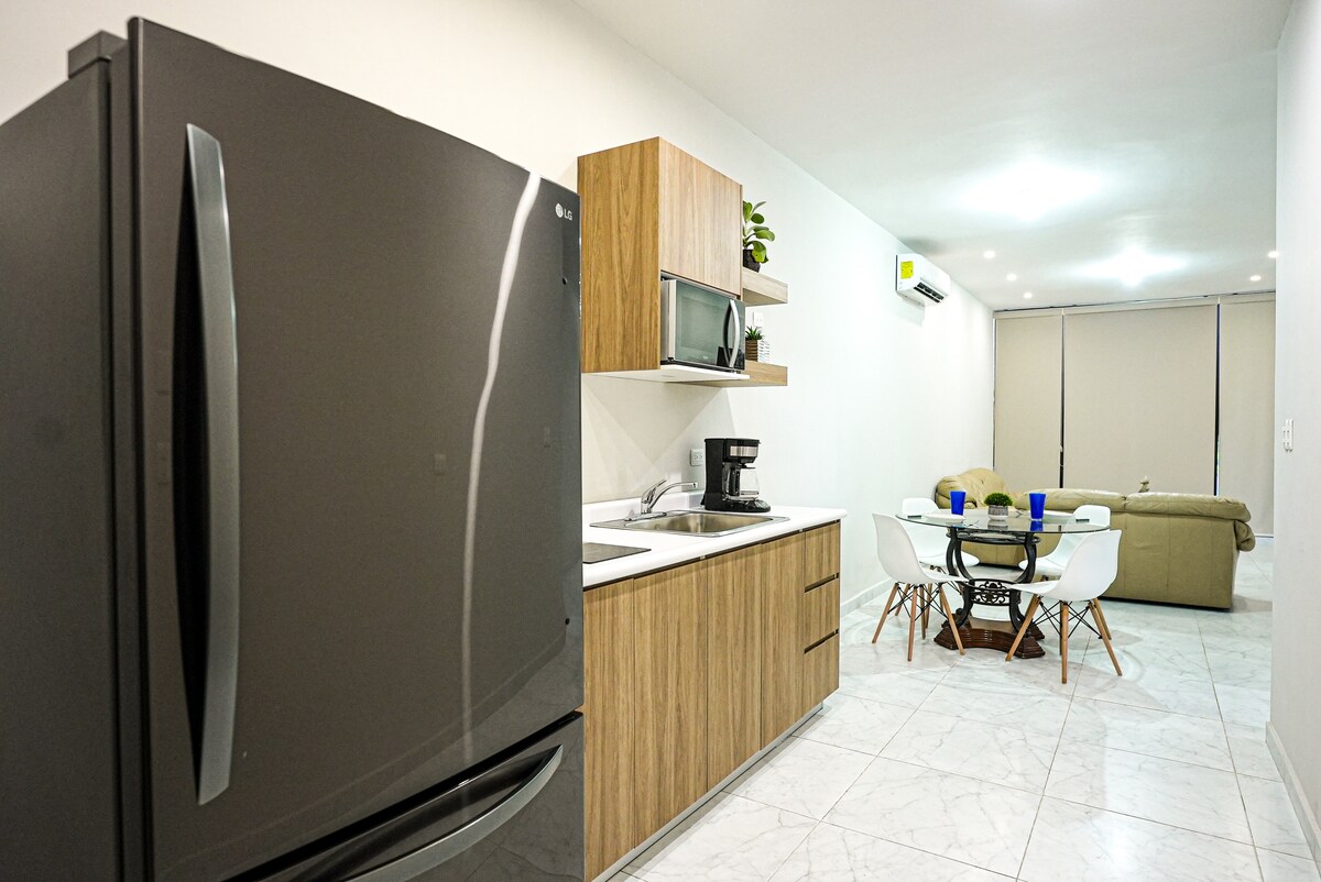 A modern kitchen area features sleek black appliances including a refrigerator, and wooden cabinetry providing ample storage. A dining space with a circular table and white chairs is visible, along with a cozy living area in the background, illuminated by soft, ambient lighting.