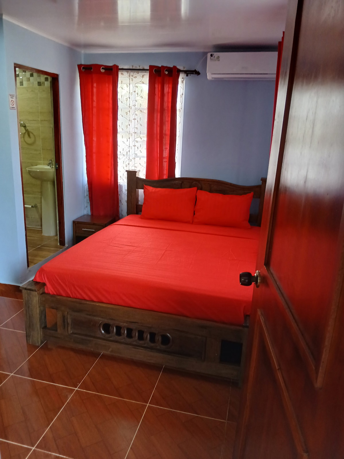 A sleeping area features a wooden bed dressed in vibrant red linens, complemented by matching pillows. Natural light filters through a window with red curtains. A nearby mirror reflects the room, while a bathroom area is visible in the background.