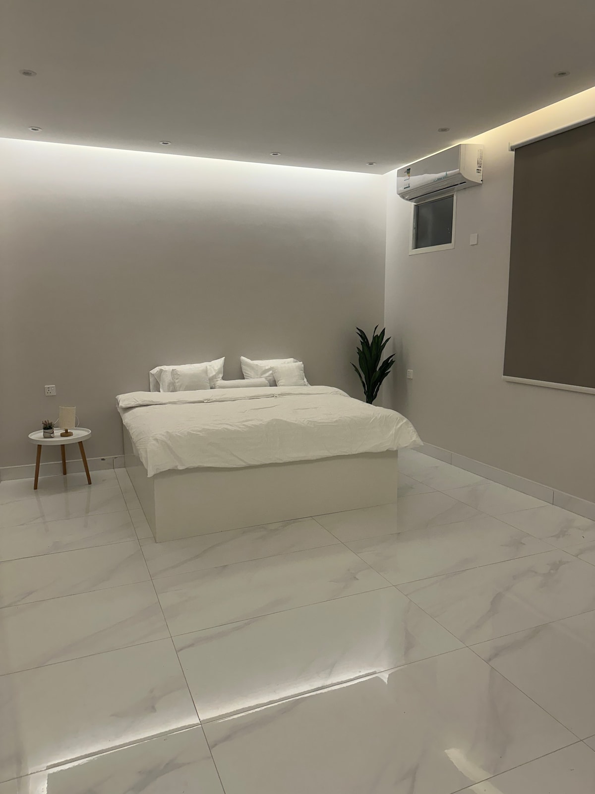A minimalist bedroom features a spacious bed adorned with white linens. A small side table holds two glasses, and a tall plant is positioned nearby. Soft lighting enhances the room’s calm ambiance, complemented by glossy marble flooring.