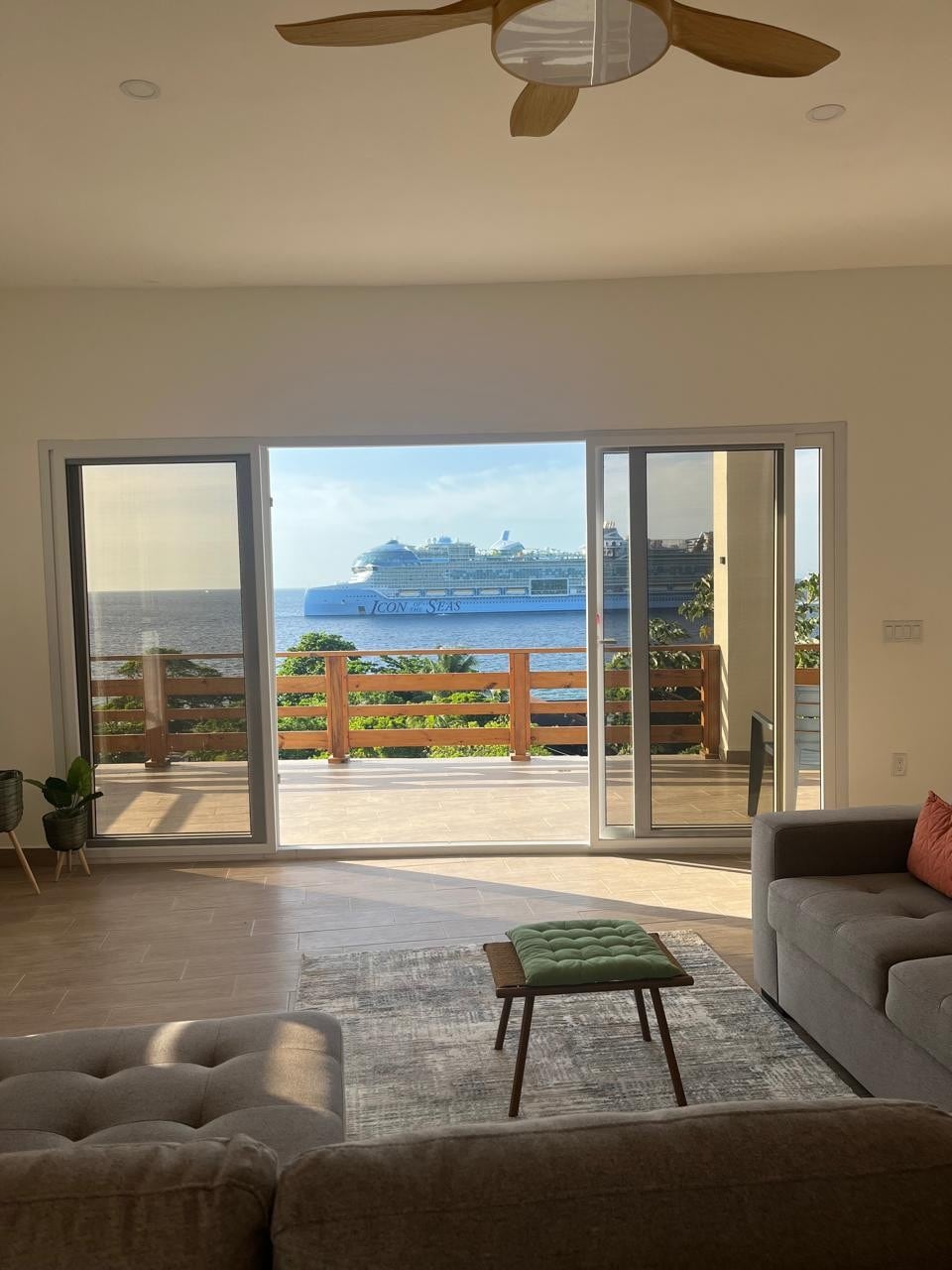 A spacious living area features large sliding glass doors, providing a view of the ocean and a cruise ship in the distance. Sunlight floods the room, highlighting a comfortable gray sofa and a green accent chair positioned on a textured rug.