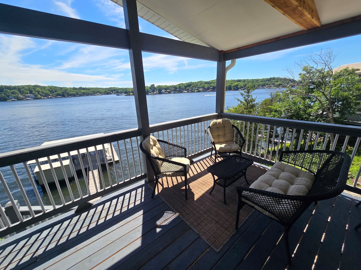 A spacious covered patio overlooks the lake, with comfortable seating arranged around a small table. The natural light illuminates the area, highlighting the view of the water and surrounding greenery. The patio provides an inviting space for relaxation and enjoying the scenery.