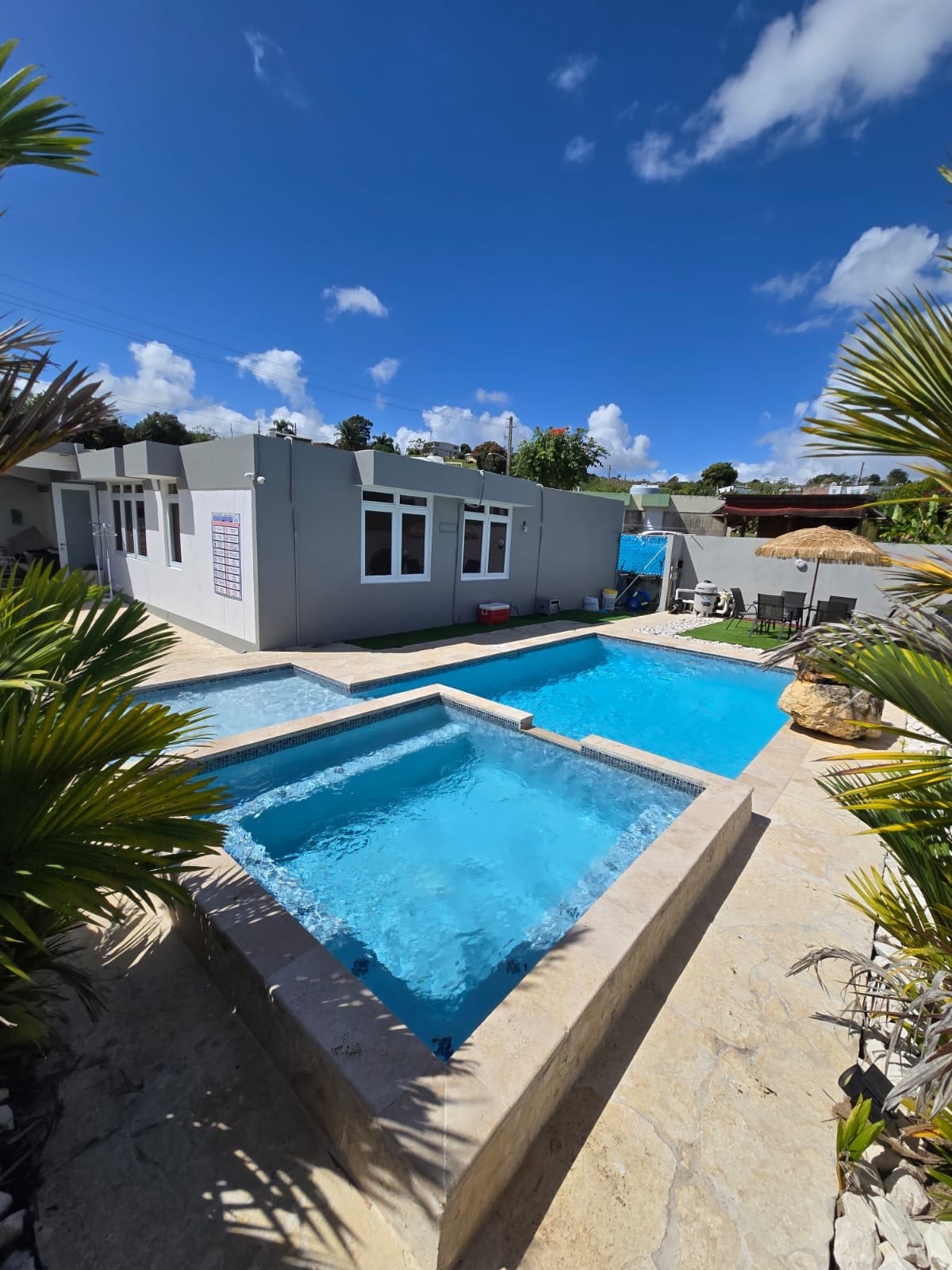 A spacious outdoor area features a pair of clean, inviting pools, surrounded by natural stone. The building layout includes multiple large windows, allowing light to fill the interior. Lush palm plants add a tropical touch, enhancing the serene environment.