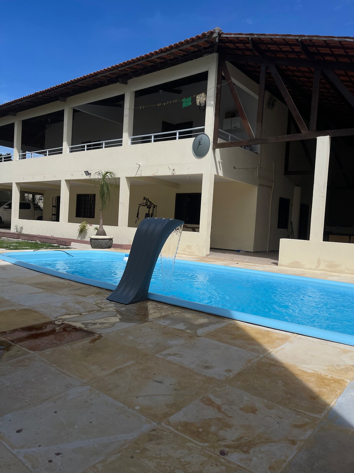 A private swimming pool is highlighted, featuring a waterfall feature that flows into the clear blue water. The surrounding area includes a spacious patio with stone flooring and a few potted plants, while the multi-story house stands in the background under a clear sky.