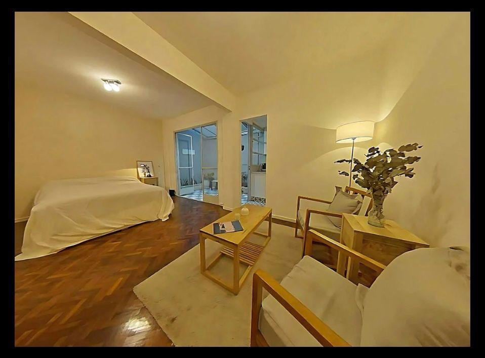 A spacious room features a large, unadorned bed covered with a light-colored blanket. Two wooden chairs are positioned next to a small coffee table, which holds a decorative vase. Soft lighting is provided by a floor lamp, illuminating the minimalist decor.