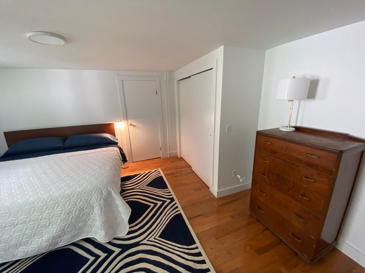 The bedroom features a spacious bed with navy bedding, complemented by a patterned rug on the hardwood floor. A tall wooden dresser is positioned beside the bed, and a lamp offers soft lighting. A closet door is visible, providing additional storage options.