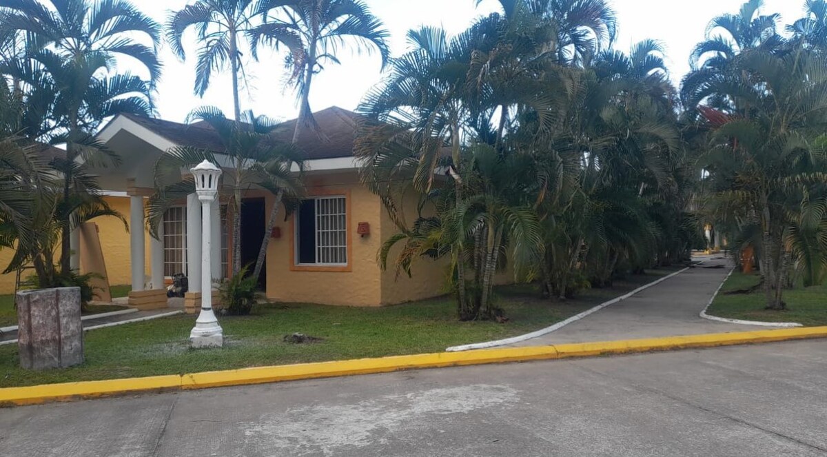 A welcoming bungalow-style villa is framed by lush tropical palm trees. The structure features a warm yellow exterior with white accents and spacious front steps leading to the entrance. A clean, paved pathway runs alongside the property, enhancing the serene outdoor setting.