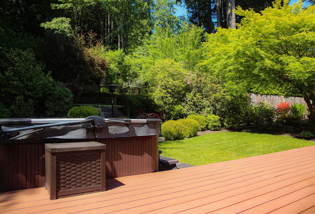 An inviting outdoor space is highlighted by a spacious deck featuring a hot tub and a decorative storage container. Lush greenery includes various trees and shrubs, creating a serene environment. The manicured garden showcases vibrant colors, adding to the natural beauty of the area.