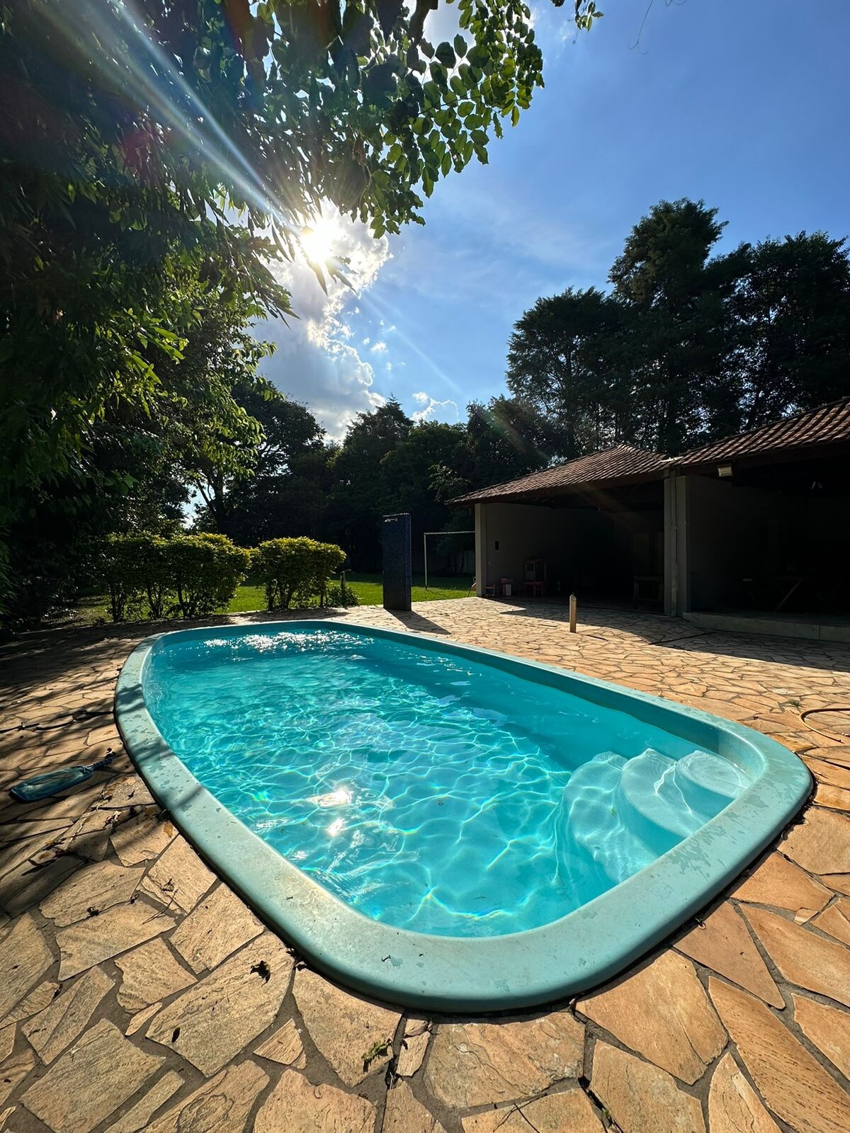 A tranquil swimming pool glistens under sunlight, framed by lush greenery and a well-manicured lawn. Natural stone flooring surrounds the pool area, offering ample space for relaxation. A covered outdoor area is visible in the background, providing shade and comfort.