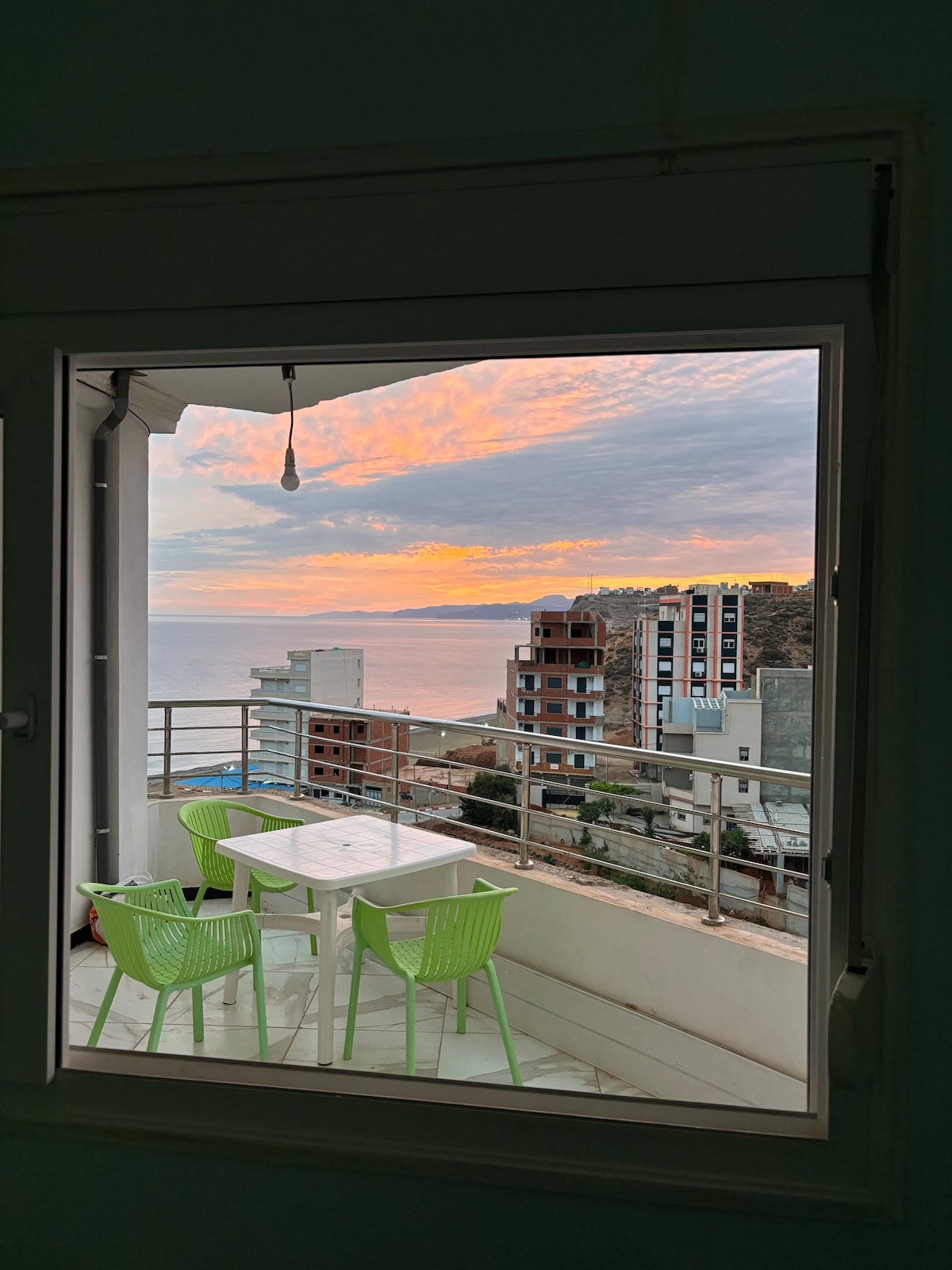 A balcony view is presented through a window, featuring a round table surrounded by green chairs. The horizon displays a soft gradient of colors as the sun sets over the ocean, with buildings visible in the foreground and background.
